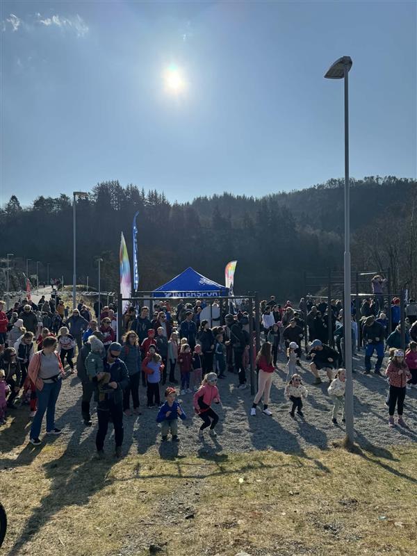 En folkemengde, deriblant barn, samles utendørs i nærheten av et blått telt på en solrik dag, med trær og åser i bakgrunnen.