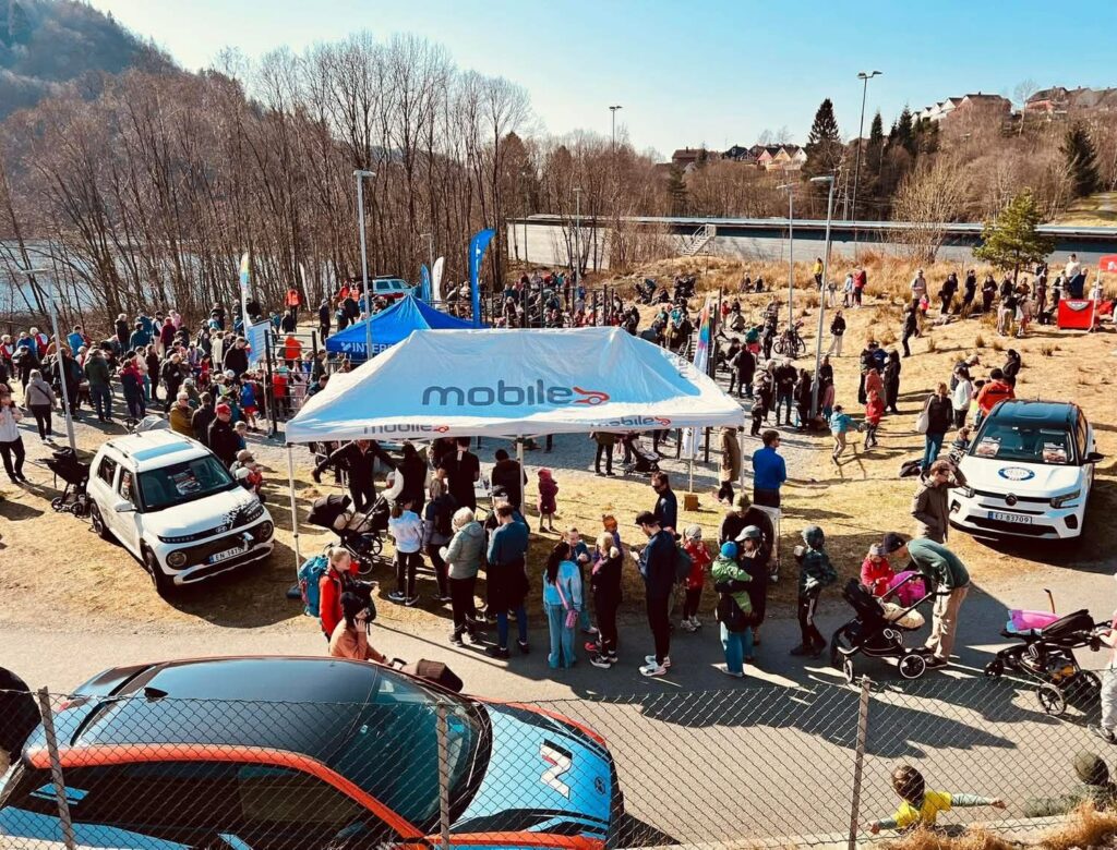 En folkemengde samles på et utendørsarrangement i nærheten av et "mobilt" telt, med parkerte biler, trær og en elv i bakgrunnen på en solfylt dag.