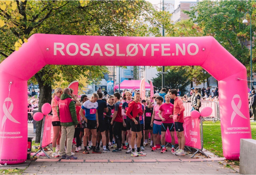 En gruppe mennesker iført rosa sportsklær samles ved startstreken under en knallrosa "ROSASLØYFE.NO"-bue på et utendørsarrangement, med trær og tilskuere i bakgrunnen.