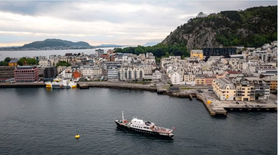 En hvit og svart båt seiler nær en kystby med lyse bygninger, en åsside med trær og en delvis overskyet himmel.