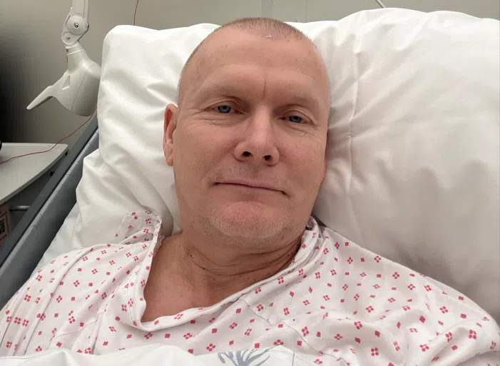 En person i en sykehusseng iført mønstret sykehuskjole ser på kameraet med et nøytralt uttrykk.