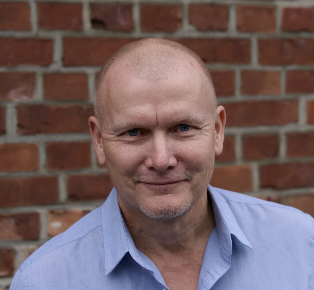 En middelaldrende mann med barbert hode og fippskjegg, iført lyseblå skjorte, står foran en rød murvegg og ser inn i kameraet.