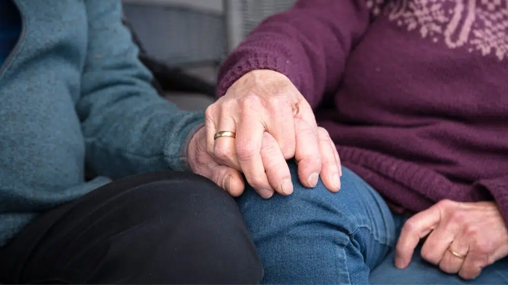 To eldre mennesker sitter side om side, den ene i blå genser og den andre i lilla, og holder hverandre i hånden på det ene benet. Begge har gifteringer.