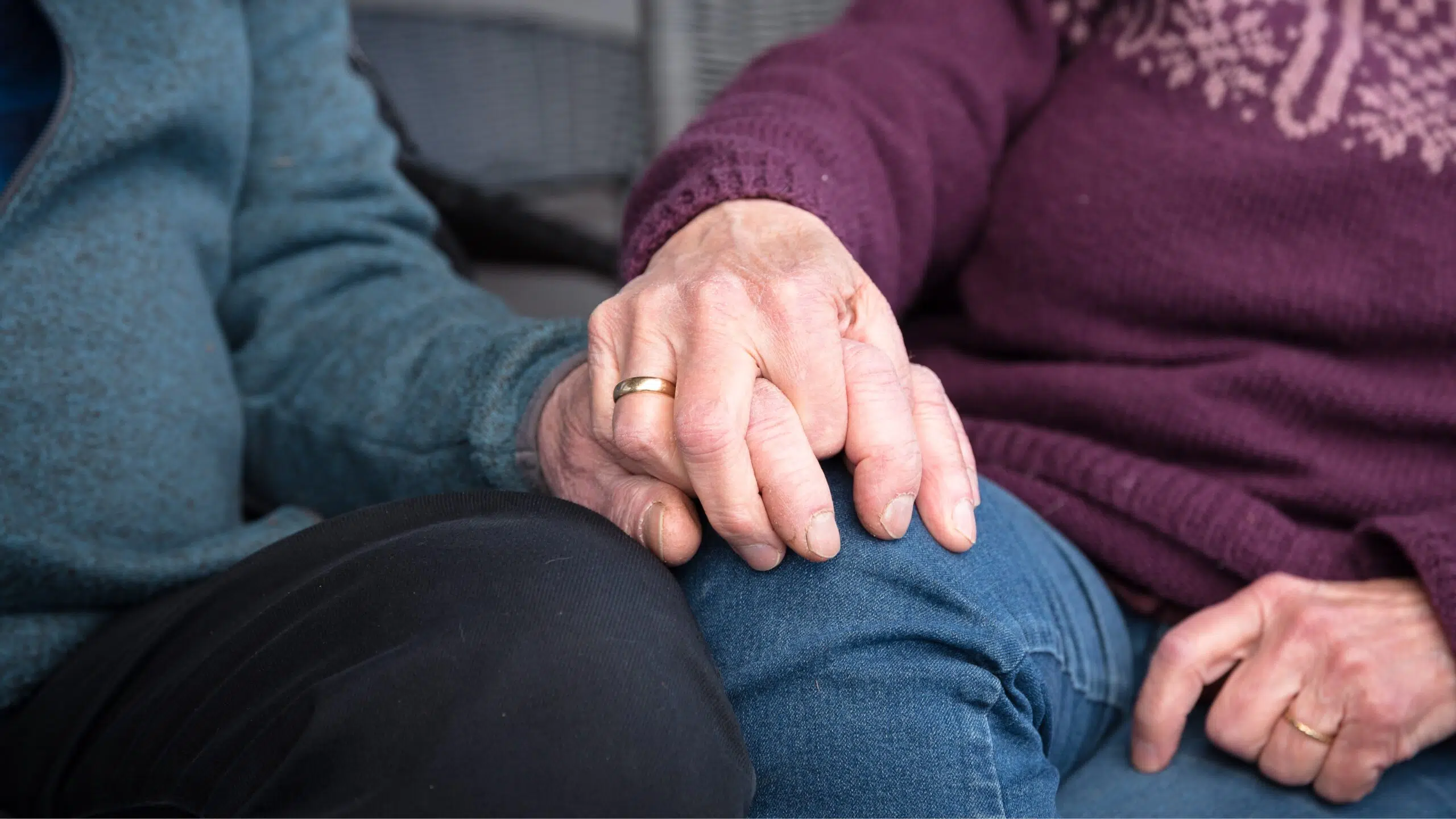 To eldre mennesker sitter side om side, den ene i blå genser og den andre i lilla, og holder hverandre i hånden på det ene benet. Begge har gifteringer.