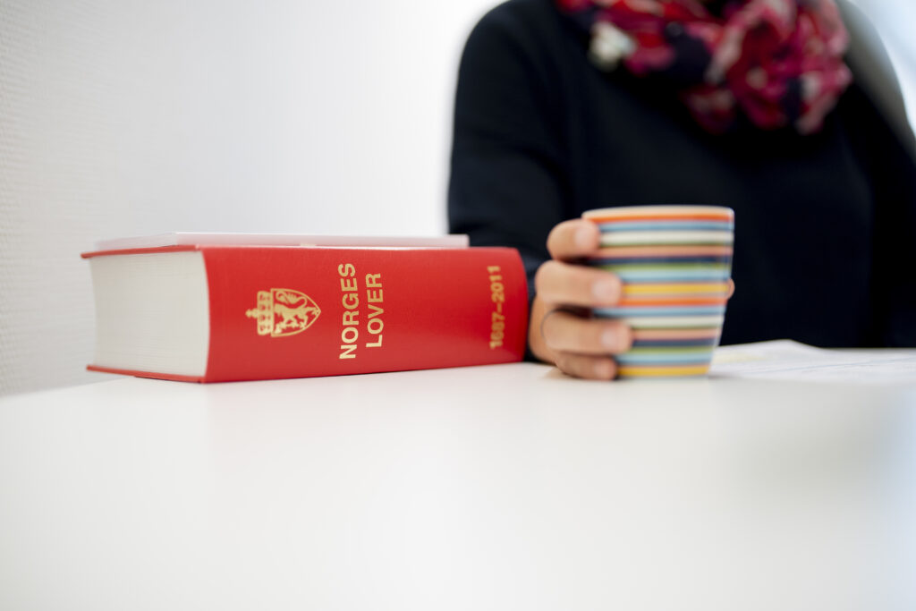 En person med en stripete kopp sitter ved et bord ved siden av en tykk, rød bok med tittelen "NORGES LOVER 1687-2011".