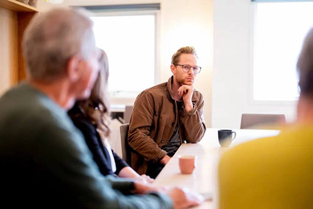 En mann med briller sitter ved et bord og hviler haken på hånden mens han lytter til andre under et møte. Flere personer er uskarpe i forgrunnen.