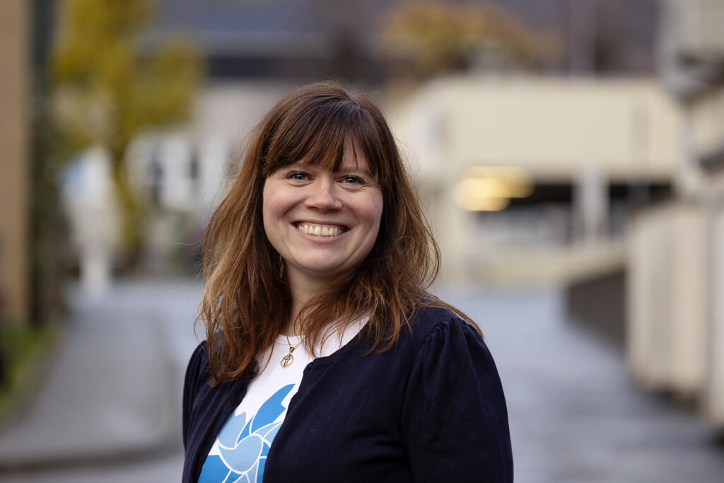 En kvinne med brunt hår og lugg, iført mørk jakke og hvit skjorte, står utendørs og smiler mot en uskarp bybakgrunn.