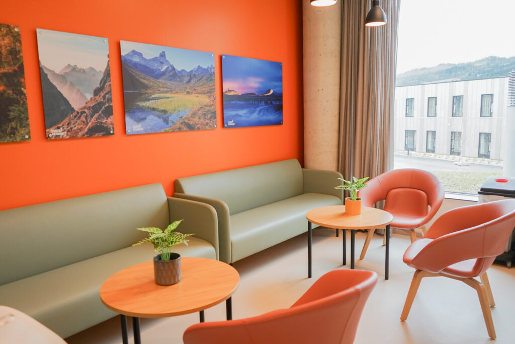 En moderne lounge med grønne sofaer, oransje stoler, runde trebord med potteplanter og landskapsbilder på en oransje vegg nær et vindu.