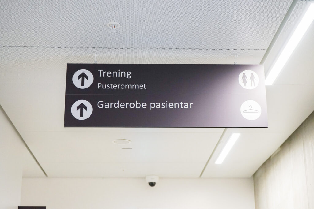 Overheadskilt med piler som viser til "Trening/Pusterommet" og "Garderobe pasientar".