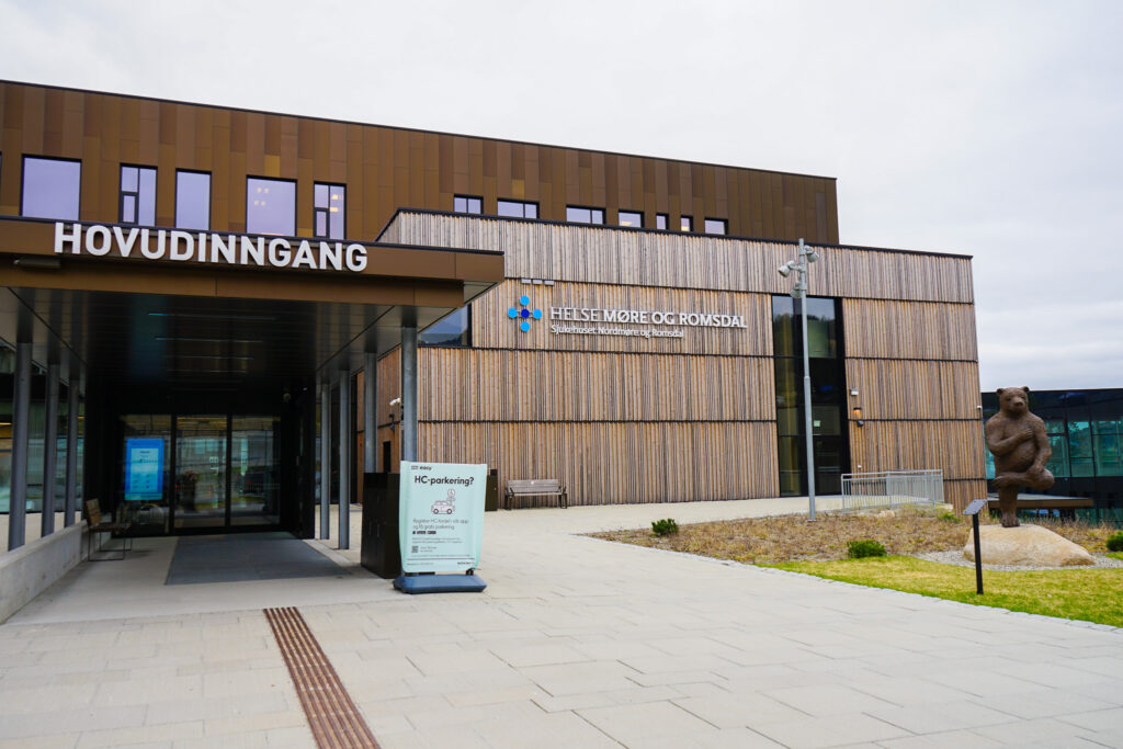 Eksteriørbilde av Helse Møre og Romsdal med hovedinngangsskilt, moderne arkitektur og en bjørnestatue til høyre.