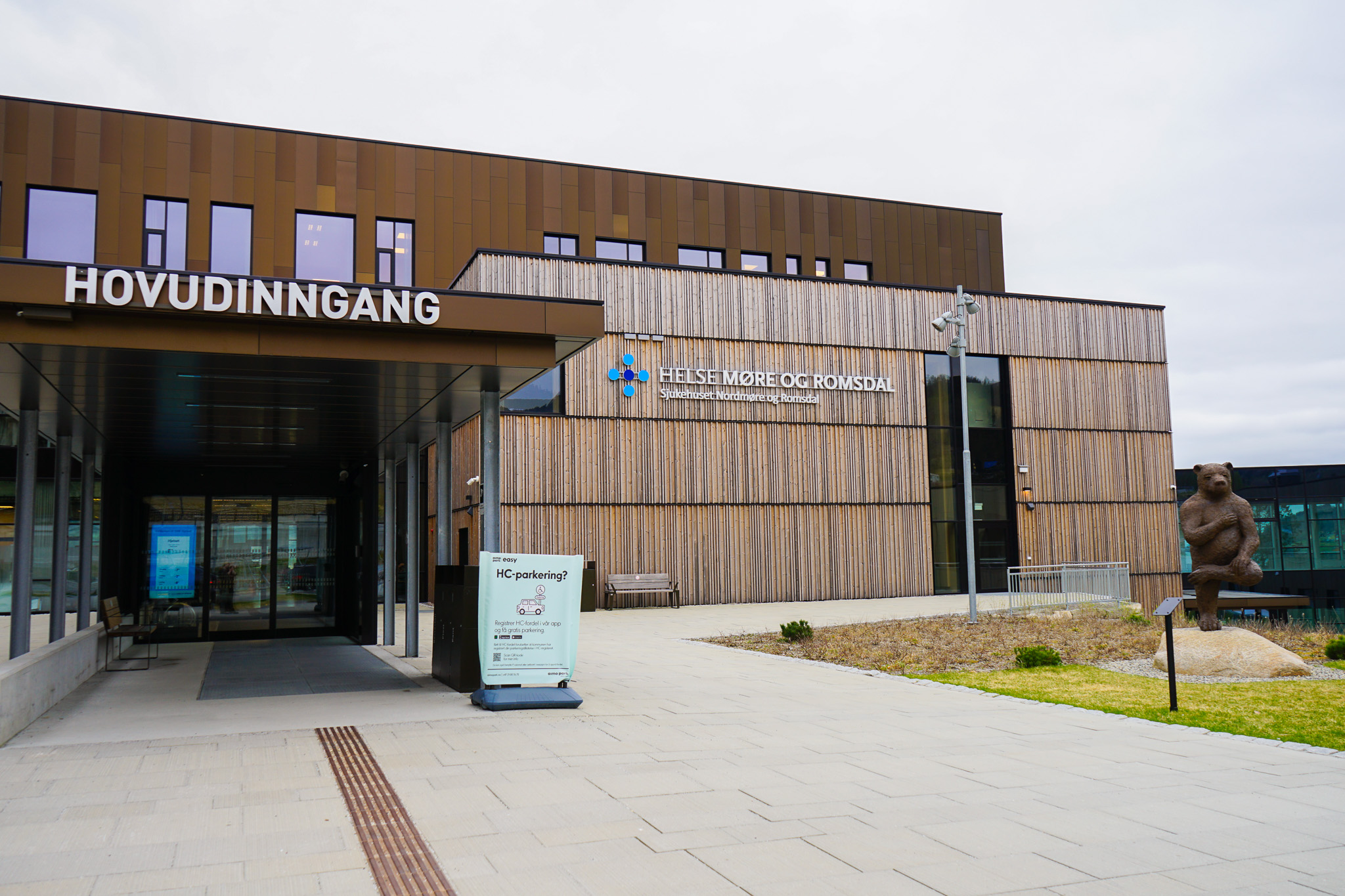 Eksteriørbilde av Helse Møre og Romsdal med hovedinngangsskilt, moderne arkitektur og en bjørnestatue til høyre.
