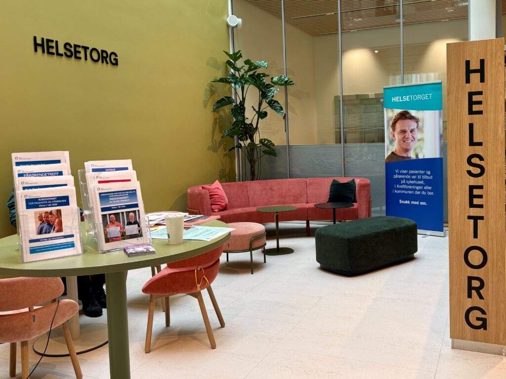 Et venteområde på Helsetorg med brosjyrer på bordene, en rosa sofa, en grønn ottoman, en potteplante og et banner med en smilende person og informasjonstekst.