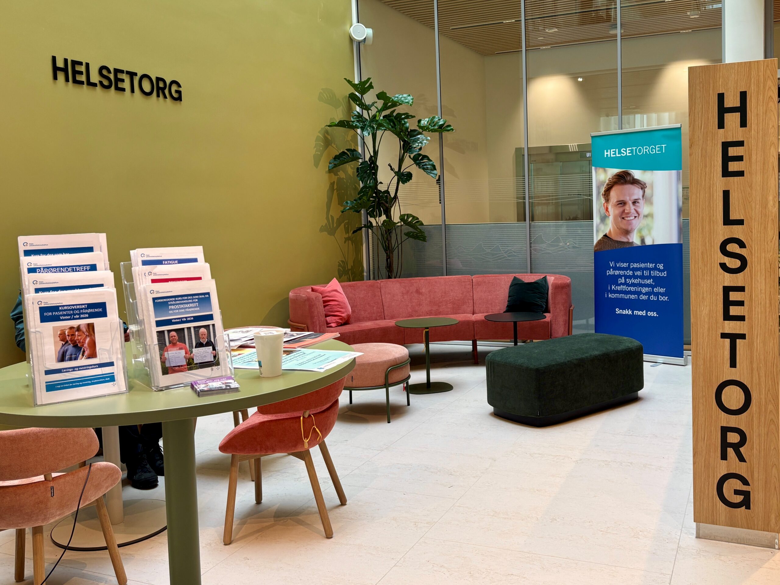 Et venteområde på Helsetorg med informasjonsbrosjyrer på bordene, en kaffekopp, en sofa, planter og et reklamebanner med en smilende person.