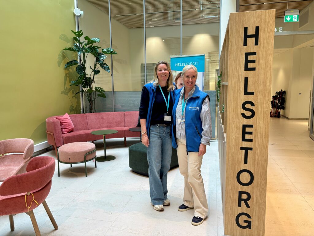 To kvinner i blå vester står og smiler i lobbyen i en moderne bygning ved siden av en trestolpe med påskriften "HELSETORG".