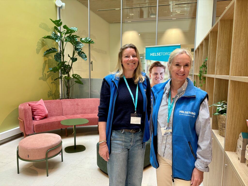 To kvinner iført blå vester og navneskilt står i en moderne lobby med en rosa sofa, en stor plante og et "HELSETORG"-banner i bakgrunnen.