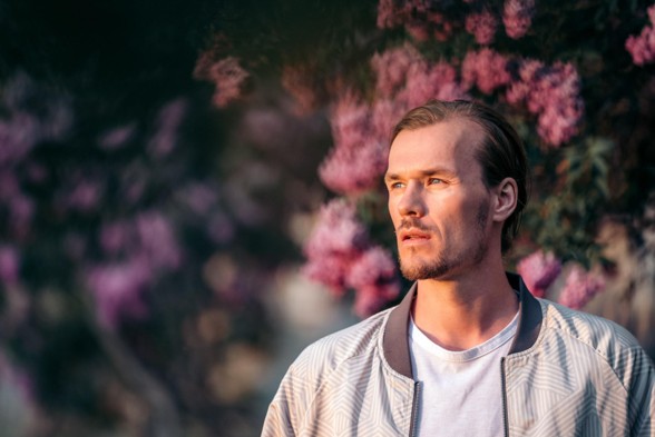 En mann med lysebrunt hår og kort skjegg står utendørs foran blomstrende rosa blomster og ser tankefullt ut i det fjerne.