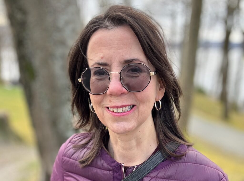 En kvinne med skulderlangt brunt hår, iført solbriller og lilla vattert jakke, står utendørs på en sti med trær og gress i bakgrunnen.