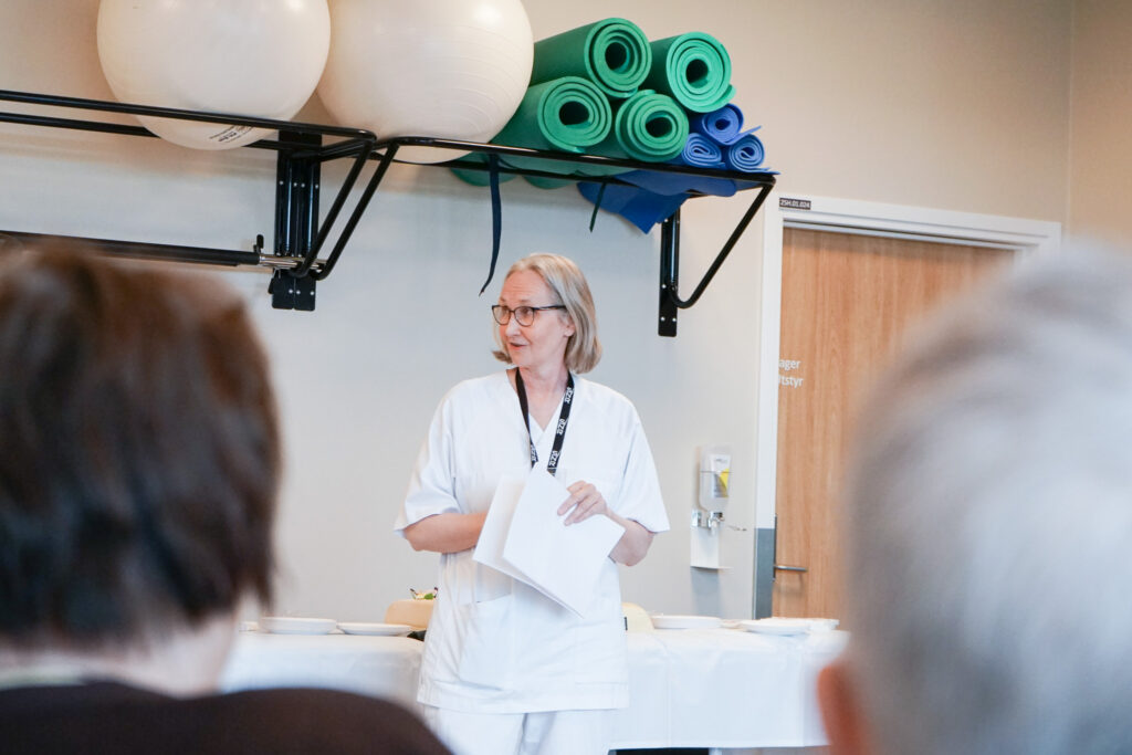 En kvinne i hvite operasjonsklær holder papirer og snakker i et rom med treningsmatter og baller på hyller bak seg, vendt mot et publikum.