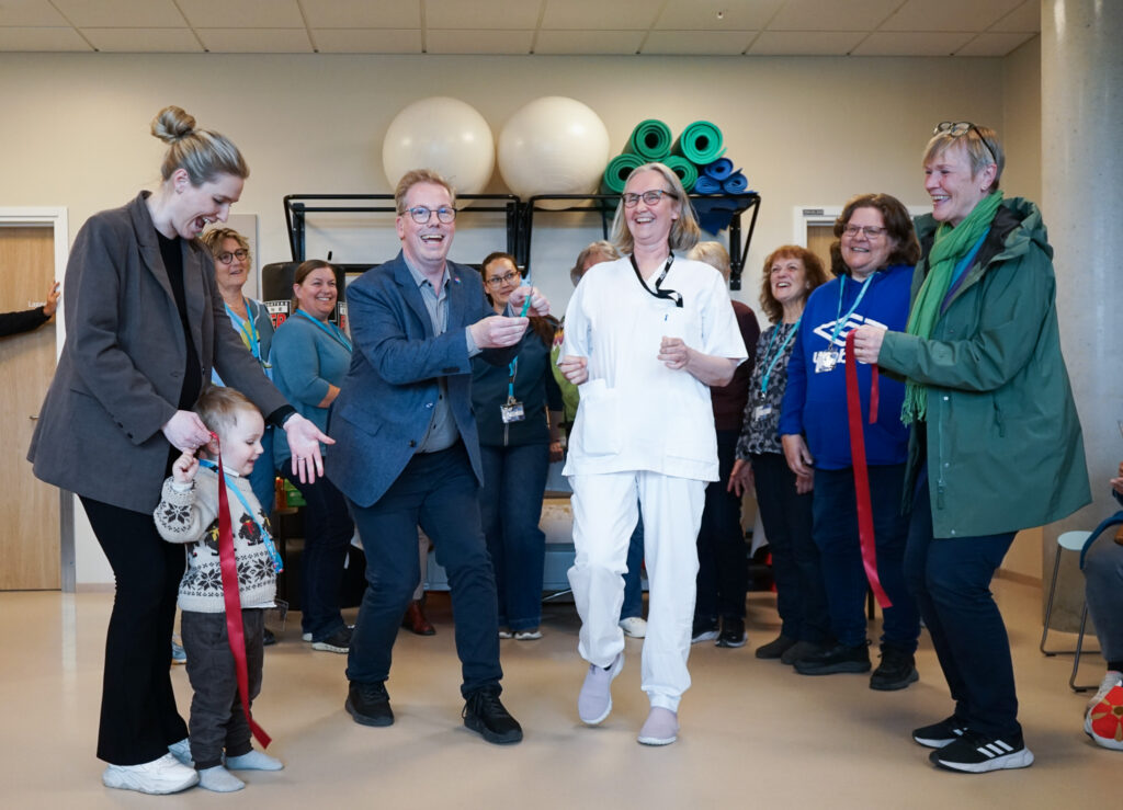 En gruppe voksne og et barn står innendørs og smiler mens en kvinne i hvite operasjonsklær klipper et rødt bånd under en seremoni.