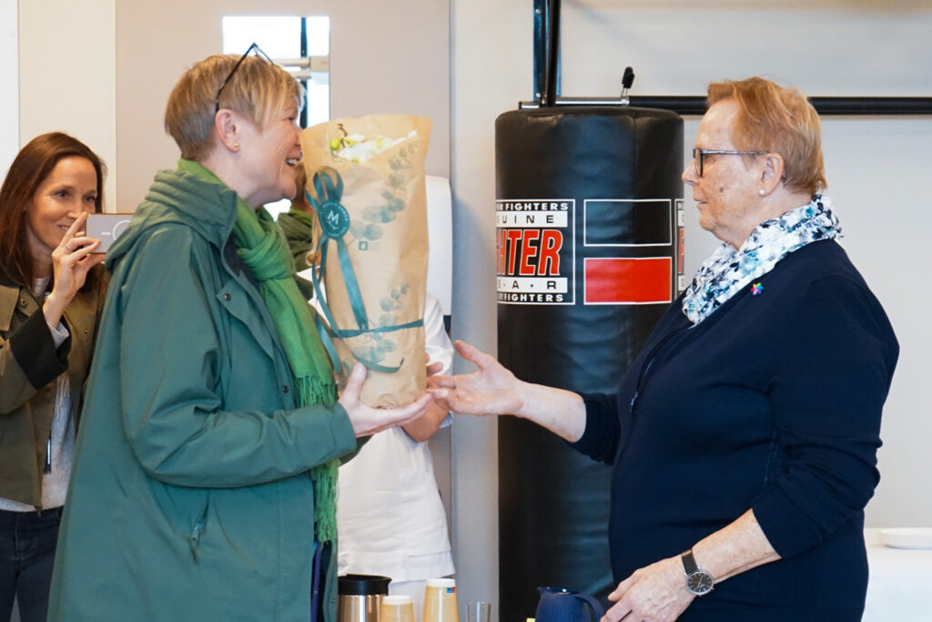 En person i grønn frakk overrekker en blomsterbukett til en eldre kvinne med briller og marineblå genser, mens andre står i bakgrunnen.