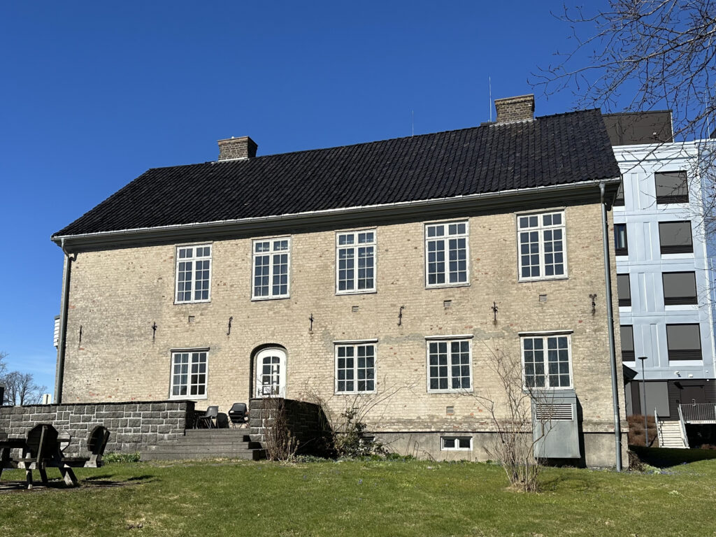 En toetasjes murbygning med svart tegltak, flere vinduer og en uteservering på en gressplen, fotografert under en klarblå himmel.