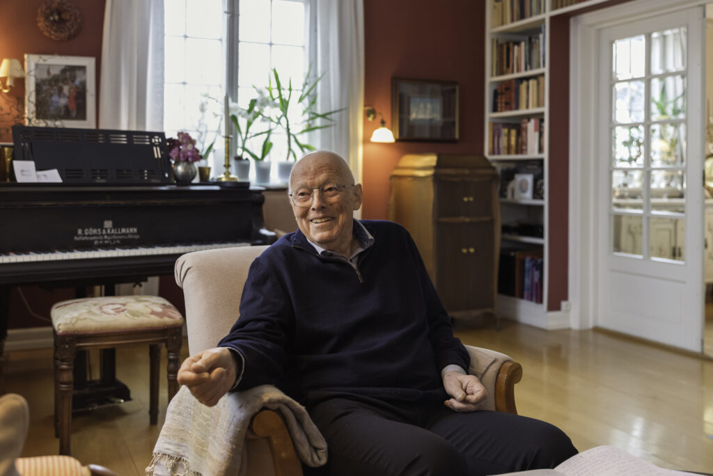 En eldre mann sitter smilende i en lenestol i en varmt opplyst stue med røde vegger, piano, bokhyller og tregulv.