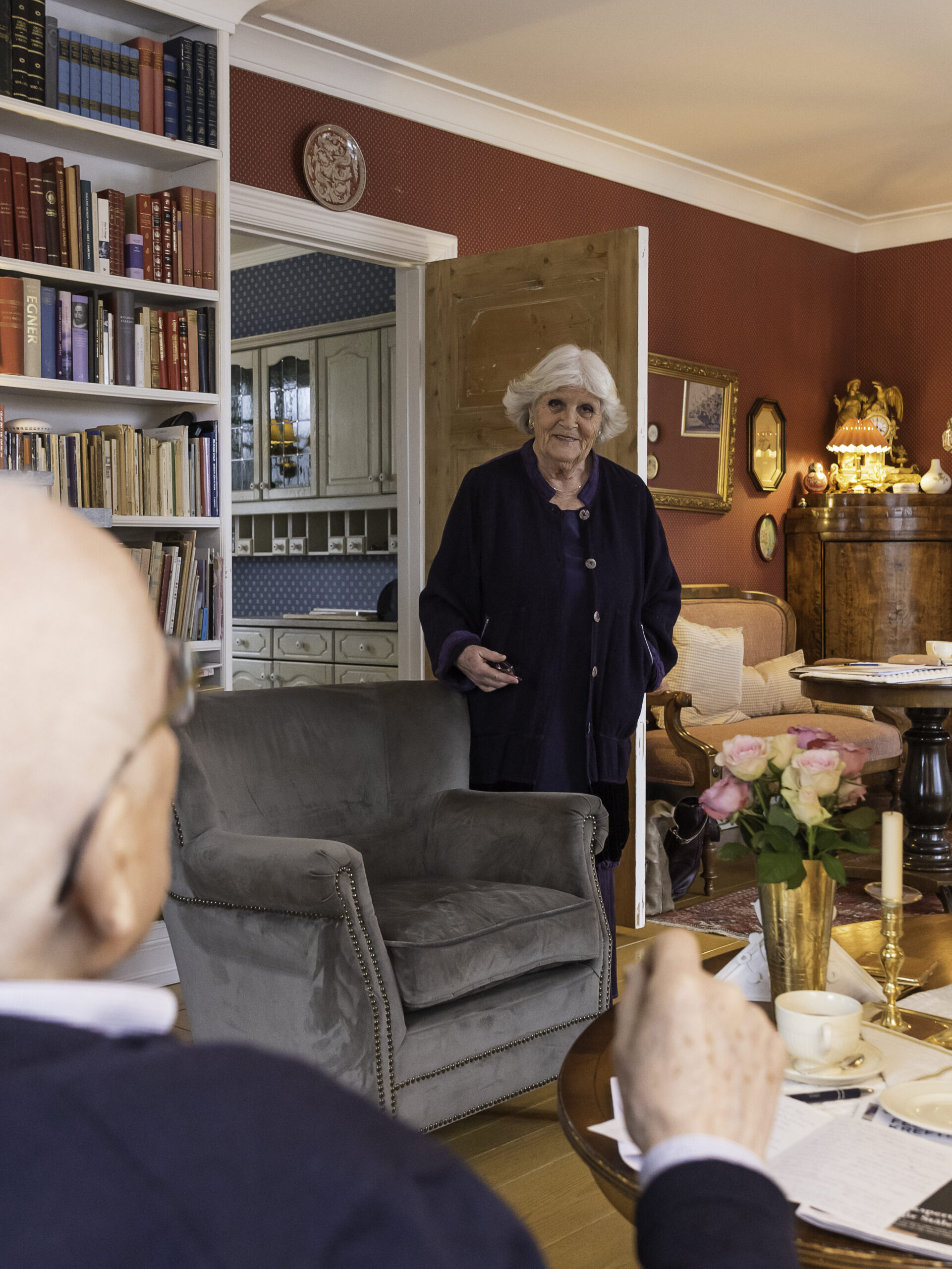 En eldre kvinne står i en koselig, bokfylt stue, vendt mot en eldre mann som sitter ved et bord dekket med kaffe, kaker og blomster.