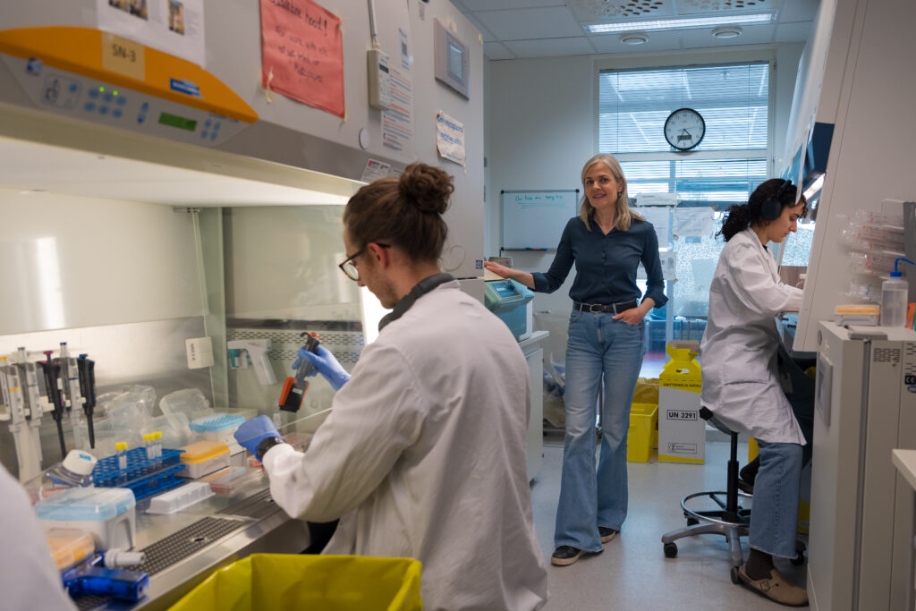 Tre personer arbeider i et laboratorium. To av dem har på seg laboratoriefrakker og håndterer utstyr, mens én person står og snakker ved en tavle.