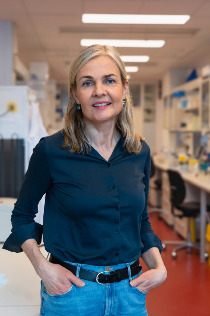 En kvinne med blondt hår, iført mørk skjorte og jeans, står i et laboratorium med hendene i lommene, vendt mot kameraet.