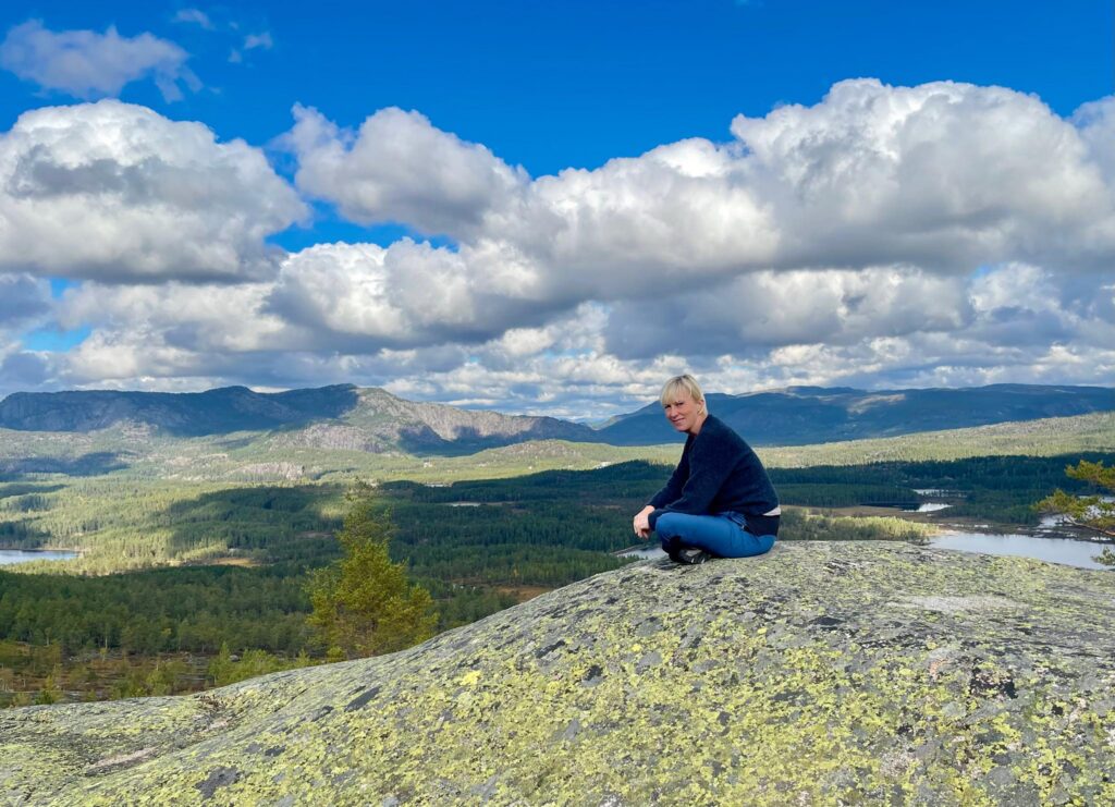 En person i mørk jakke og blå bukser sitter på en steinete bakketopp med utsikt over et vidstrakt skoglandskap under en delvis overskyet himmel.