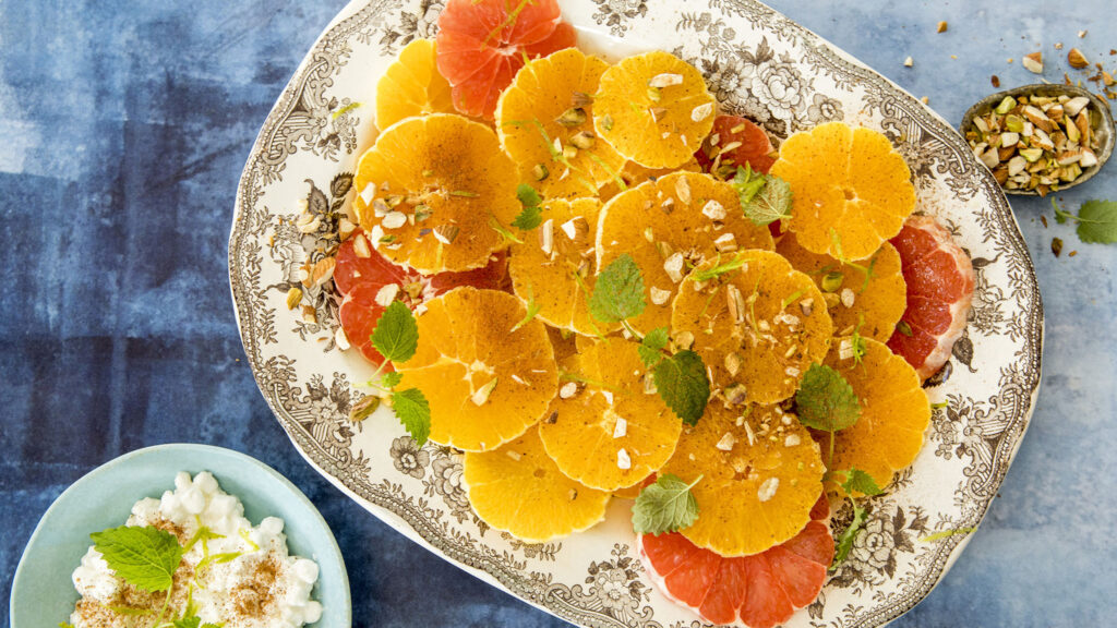 Et fat med appelsiner og grapefrukt i skiver, pyntet med mynteblader og hakkede nøtter på et mønstret fat, og en bolle med cottage cheese ved siden av.