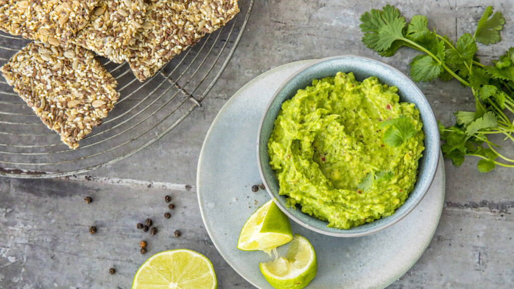 En bolle med guacamole pyntet med koriander, servert med limebåter på en tallerken, ved siden av frøkjeks og fersk koriander på en grå flate.
