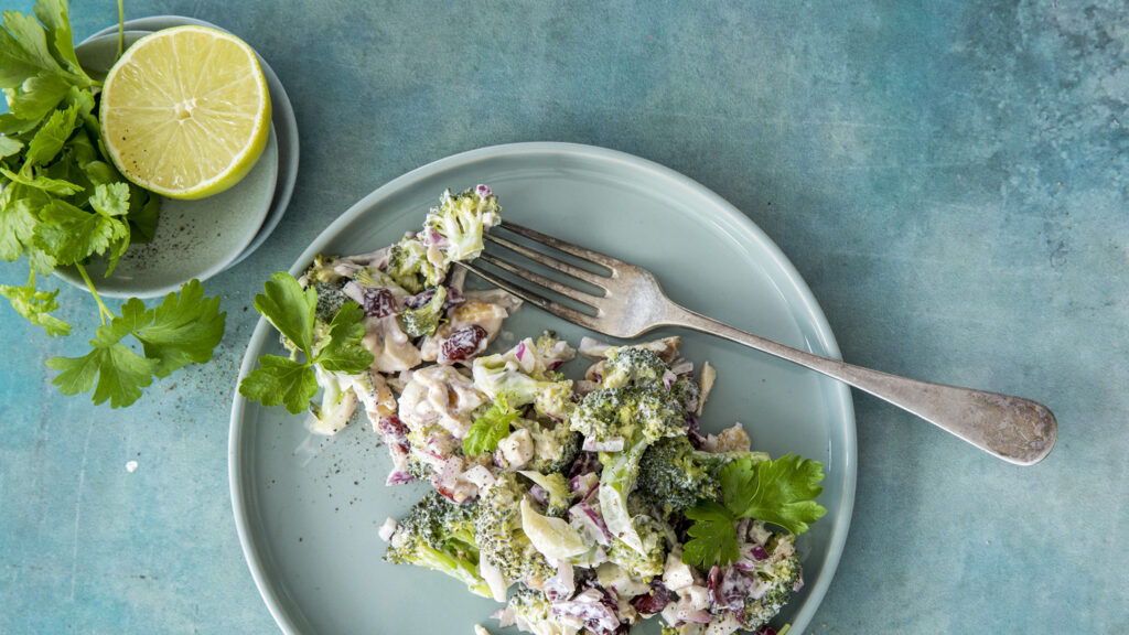 En tallerken med brokkolisalat, persillegarnityr og en gaffel ved siden av en liten bolle med en halv lime og fersk persille på blå bakgrunn.