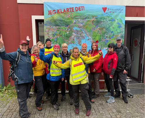 En gruppe mennesker i friluftsutstyr poserer og smiler foran et stort, fargerikt kart med teksten "JEG KLARTE DET!" trykket øverst.