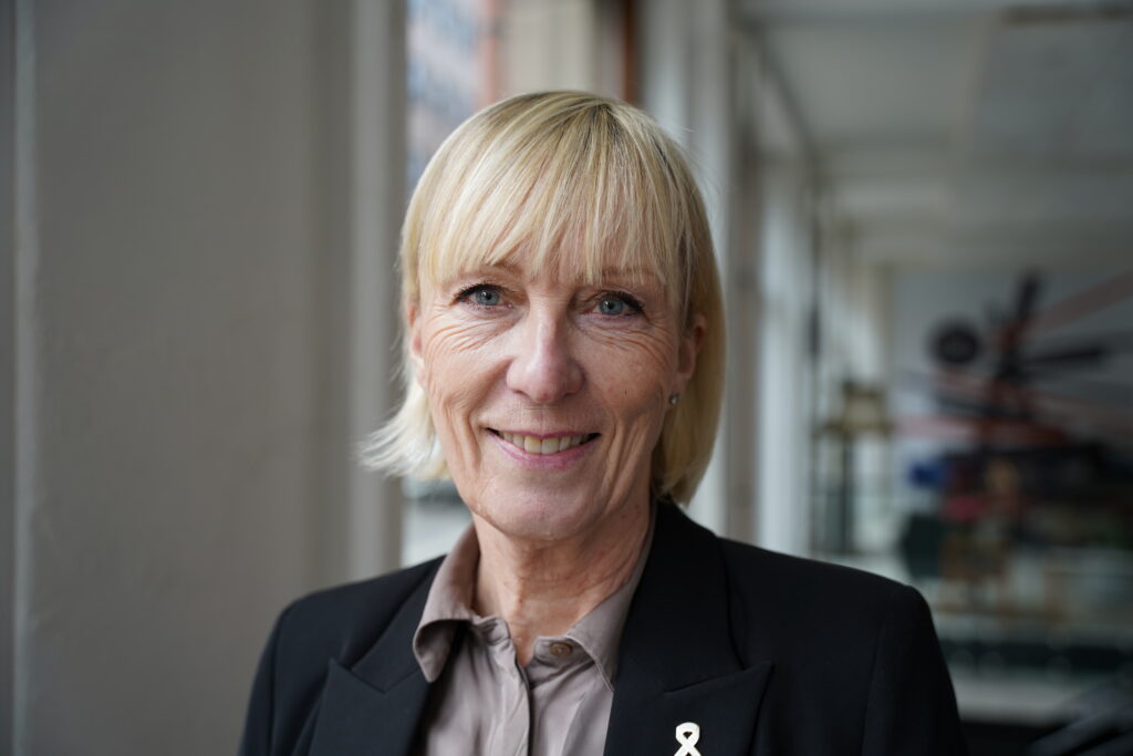 En kvinne med kort, blondt hår og svart blazer smiler til kameraet i en lys innendørs gang.