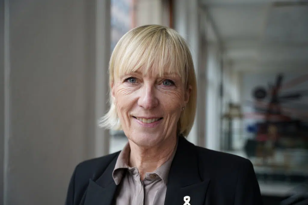 En kvinne med kort, blondt hår, iført svart blazer og beige bluse, står innendørs og smiler til kameraet.