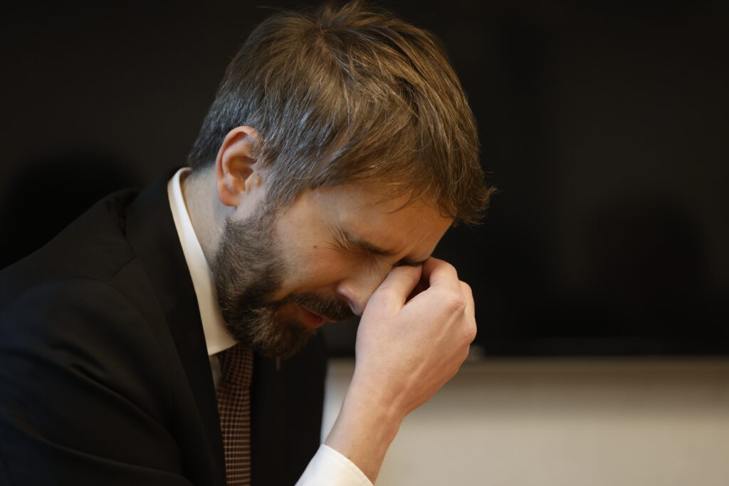 En mann i dress sitter innendørs med lukkede øyne og gnir seg på neseryggen med hånden, og virker stresset eller trett.