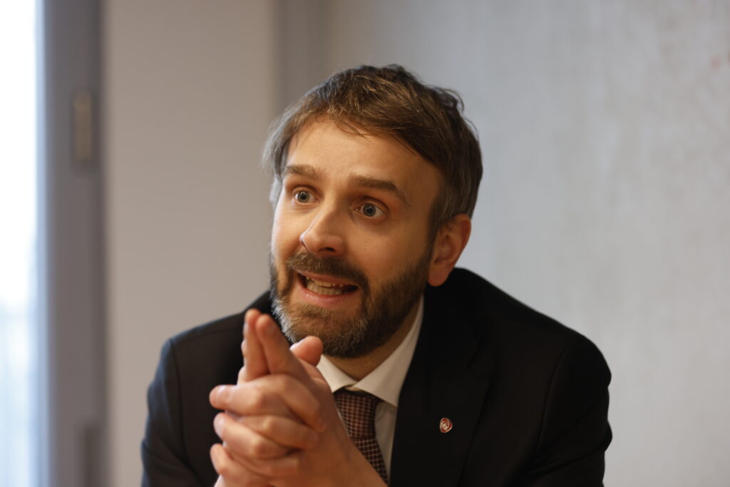 En mann i dress sitter ved et bord, knytter hendene og snakker med et uttrykksfullt ansiktsuttrykk.
