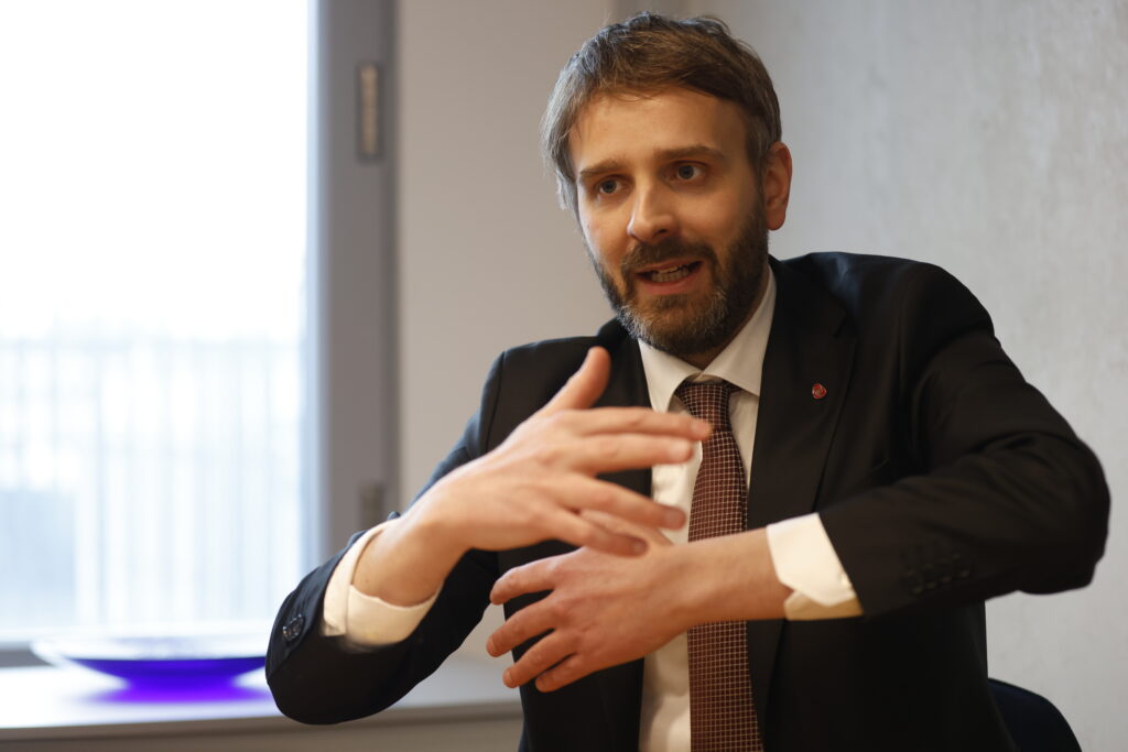En mann i dress og slips gestikulerer med hendene mens han snakker, sittende innendørs ved et vindu.