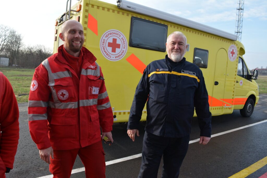 To menn, en i rød legeuniform og en i mørk uniform, står smilende foran en gul ambulanse med Røde Kors-logoer på en asfaltert vei.