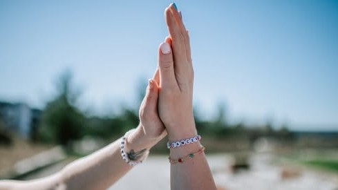 To hender med armbånd som gir hverandre en high five utendørs, med klar blå himmel og uskarp bakgrunn.