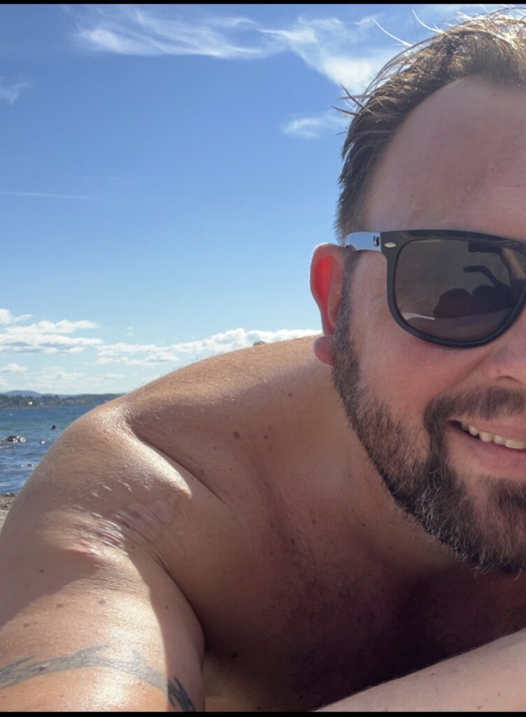 En mann med skjegg og solbriller ligger på en strand under en blå himmel, med vann og fjerne mennesker i bakgrunnen.