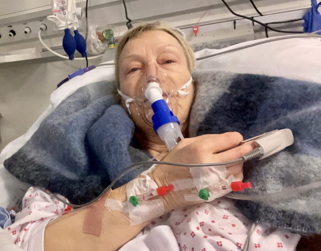 En eldre pasient ligger i en sykehusseng med oksygenmaske, intravenøs slange og overvåkingsutstyr festet til hånden, pakket inn i et teppe.