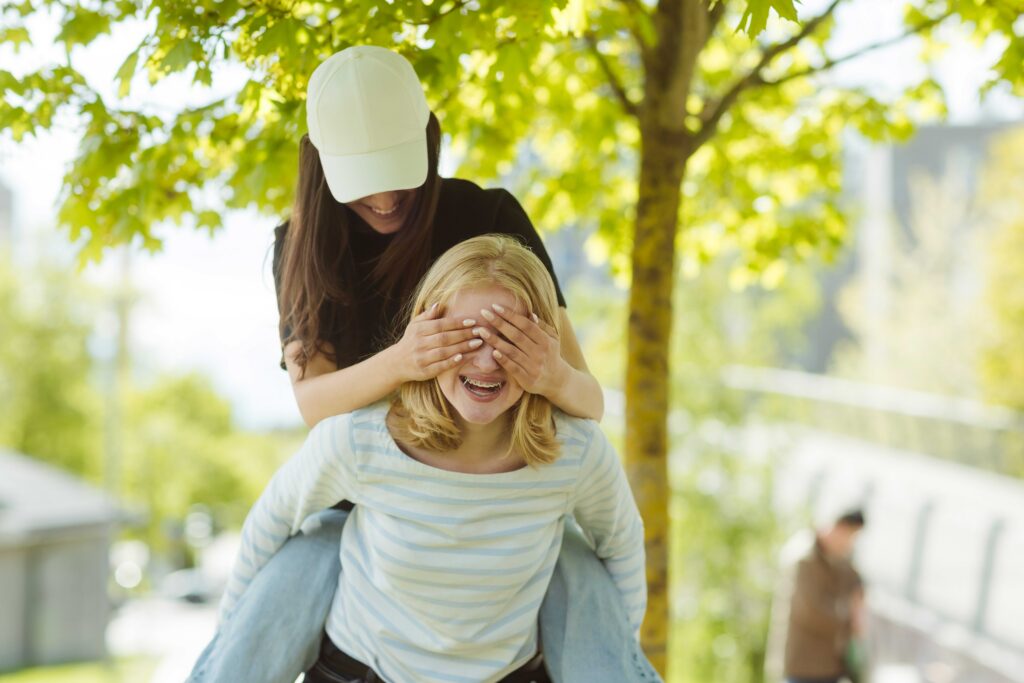 En person med hette dekker øynene til en annen person som smiler mens han eller hun gir dem en ridetur utendørs under et tre.