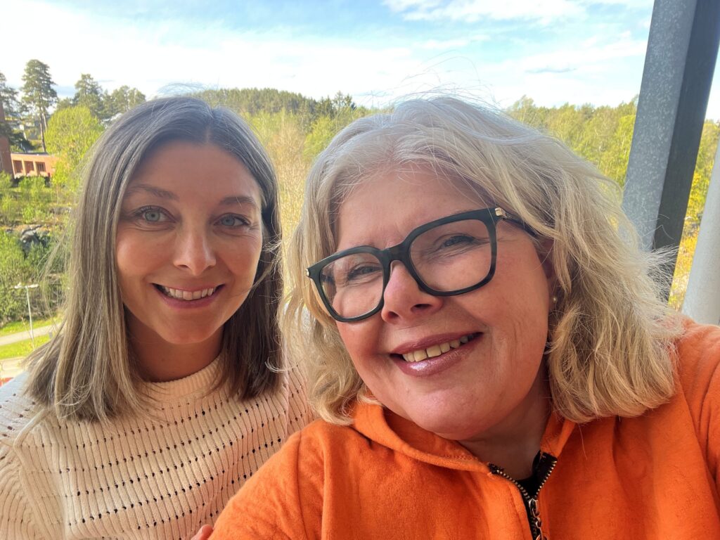 To kvinner smiler utendørs, den ene med briller og oransje topp, den andre i hvit genser, med grønne trær og blå himmel i bakgrunnen.