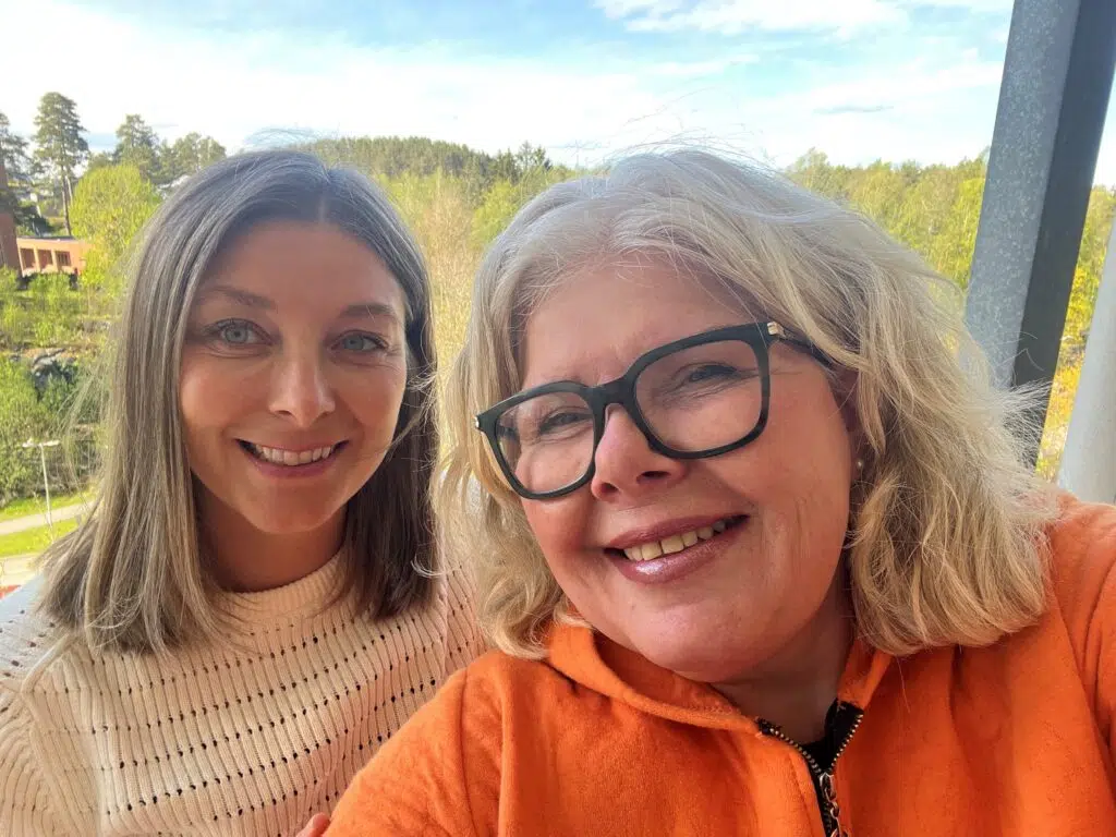 To kvinner smiler utendørs, den ene med briller og oransje topp, den andre i hvit genser, med grønne trær og blå himmel i bakgrunnen.