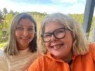 To kvinner smiler utendørs, den ene med briller og oransje topp, den andre i hvit genser, med grønne trær og blå himmel i bakgrunnen.