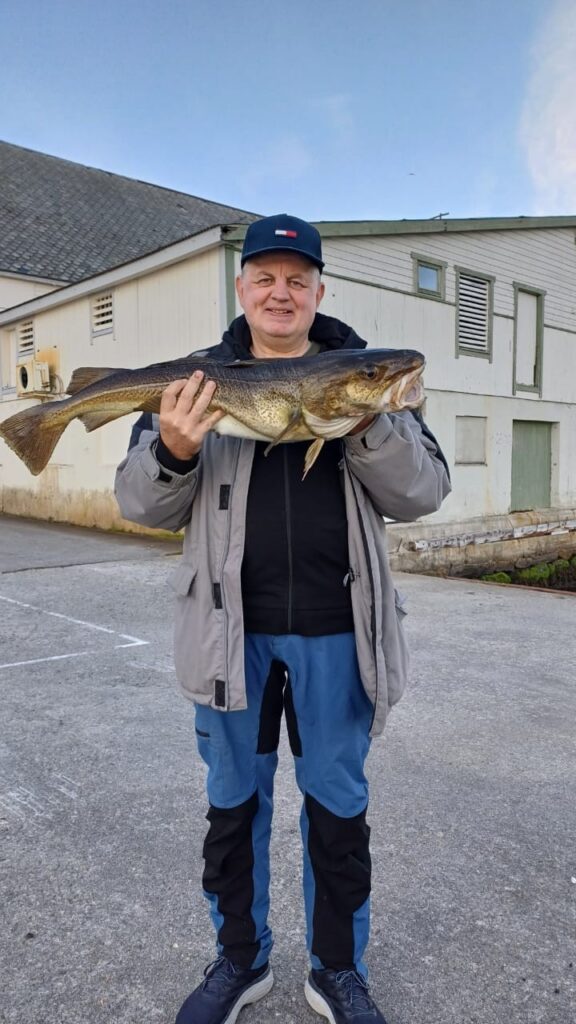 En mann står utendørs på betong og holder en stor fisk med begge hender, foran en hvit bygning med grønne dører og vinduer.