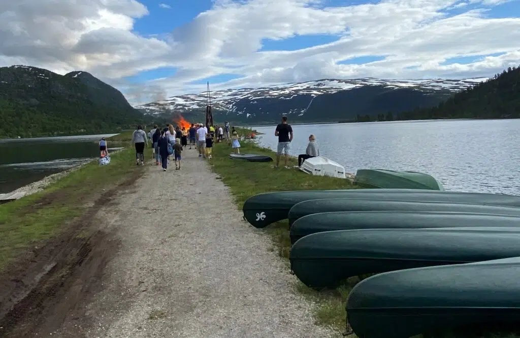 Folk går langs en sti ved innsjøen mot et stort bål, med flere grønne kanoer som ligger på gresset og snødekte fjell i bakgrunnen.