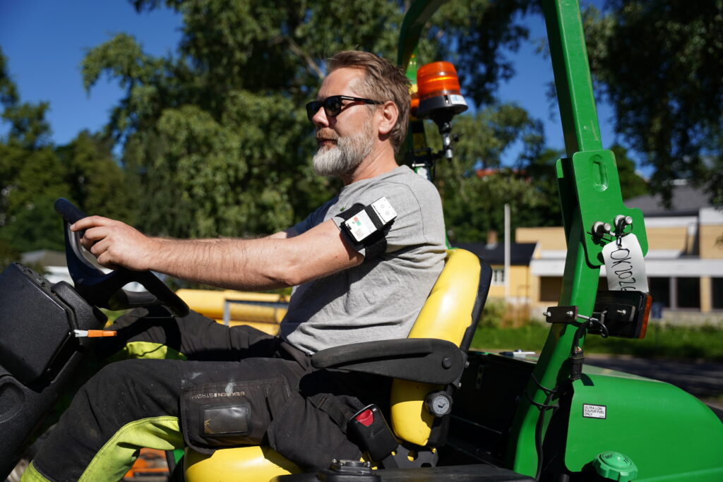 En mann med grått skjegg sitter på et grønt arbeidsbil, iført solbriller og med en overvåkingsenhet på overarmen, utendørs på en solrik dag.
